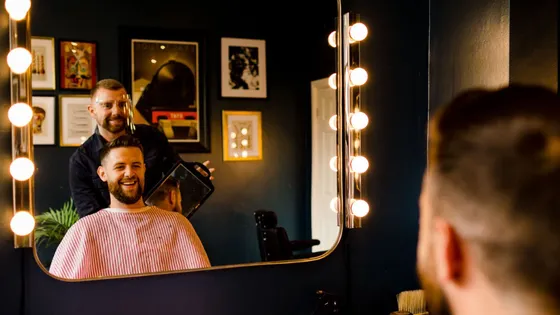 A bearded client in the chair at Butler’s Barbering, navy walls and bulb-lit mirrors behind